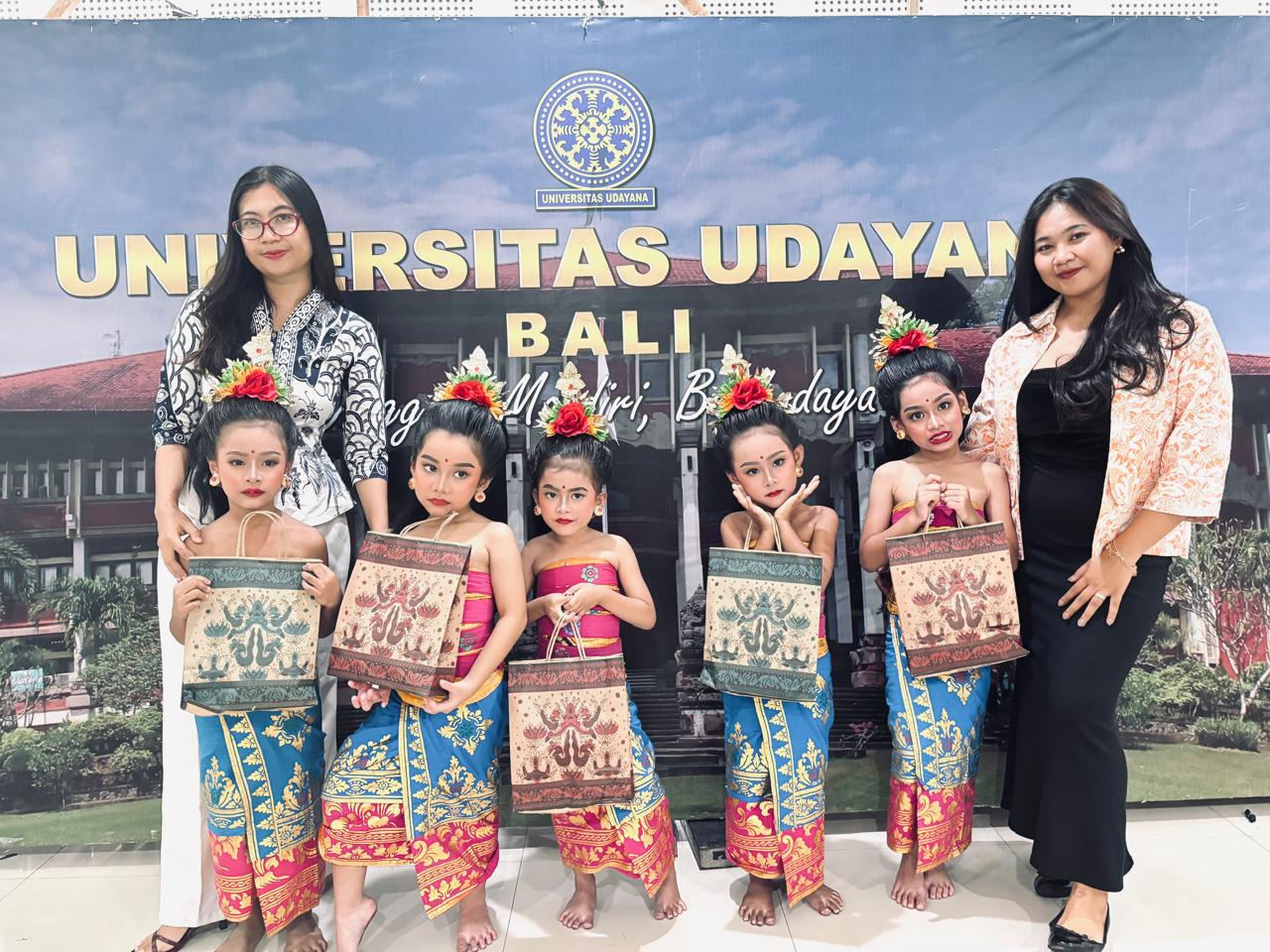 Two Balinese educators with five young students in traditional ceremonial dress and floral headpieces at Universitas Udayana, Bali